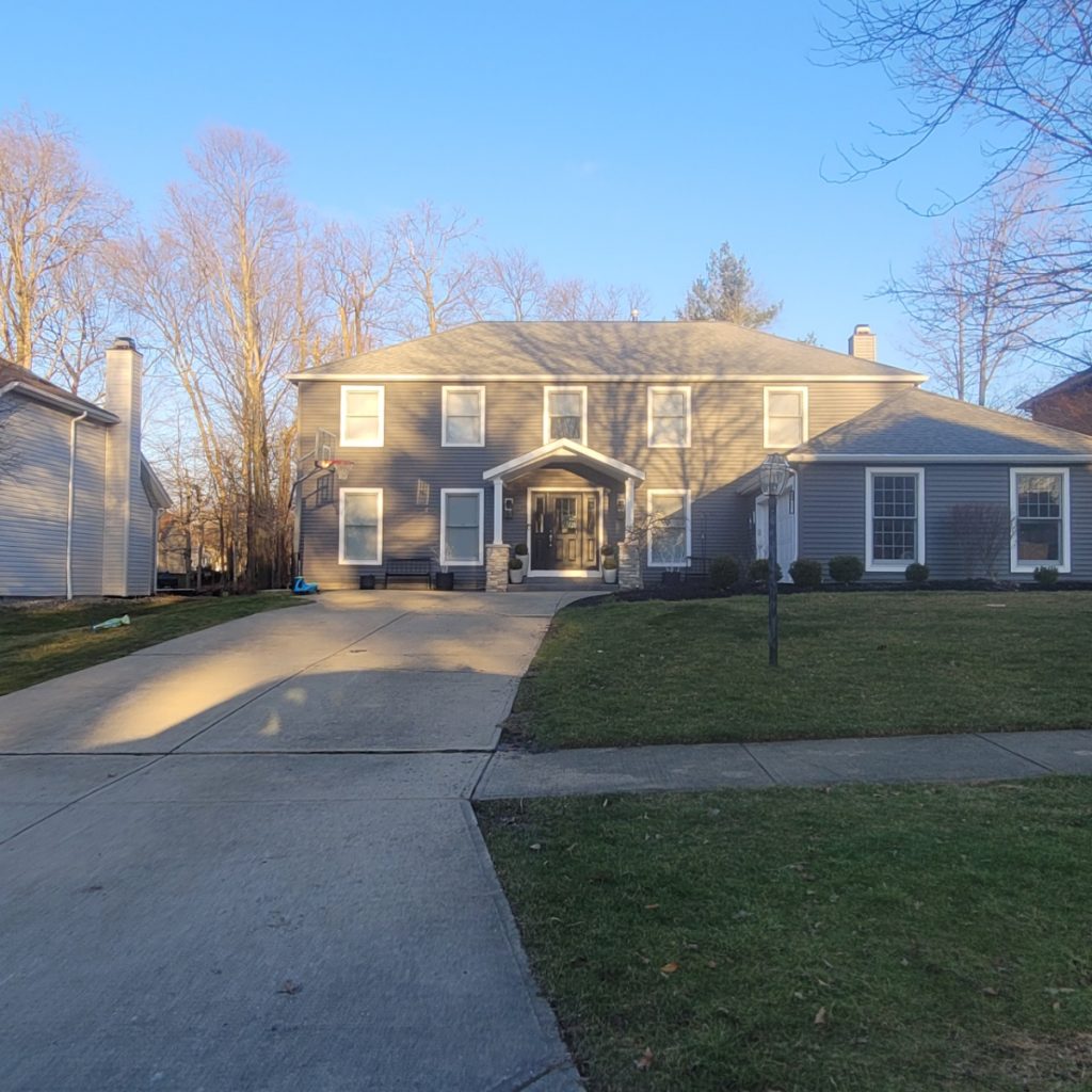 A two-story colonial-style house with light gray vinyl siding is bathed in the cool light of a clear day. A paved driveway leads to the front of the house, which features a covered entryway with columns. Green grass lawns are visible in the foreground on either side of the driveway and a sidewalk runs along the edge of the property. Bare trees are visible in the background, suggesting late autumn or winter. The house has multiple windows with white trim.