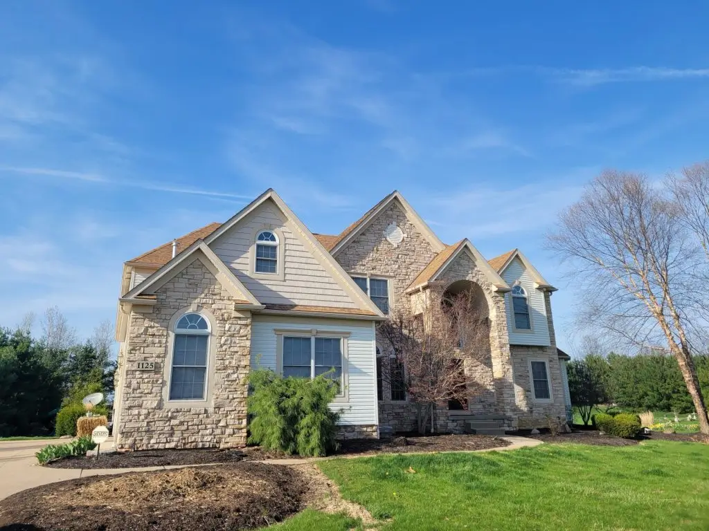Split-level home with white siding, black shutters, and a blue front door, set on a grassy lawn with a brick foundation and autumn trees in the background.