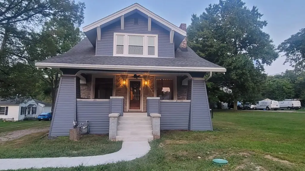 A two-story colonial-style house with light gray vinyl siding is bathed in the cool light of a clear day. A paved driveway leads to the front of the house, which features a covered entryway with columns. Green grass lawns are visible in the foreground on either side of the driveway and a sidewalk runs along the edge of the property. Bare trees are visible in the background, suggesting late autumn or winter. The house has multiple windows with white trim.