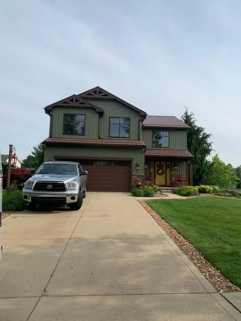 Two-story colonial-style home with sage green vinyl siding, white trim, black-framed windows, and a dark gray asphalt shingle roof. Features a black front door with sidelights, a small wooden porch with natural railing, and neatly landscaped flower beds with shrubs and spring flowers.