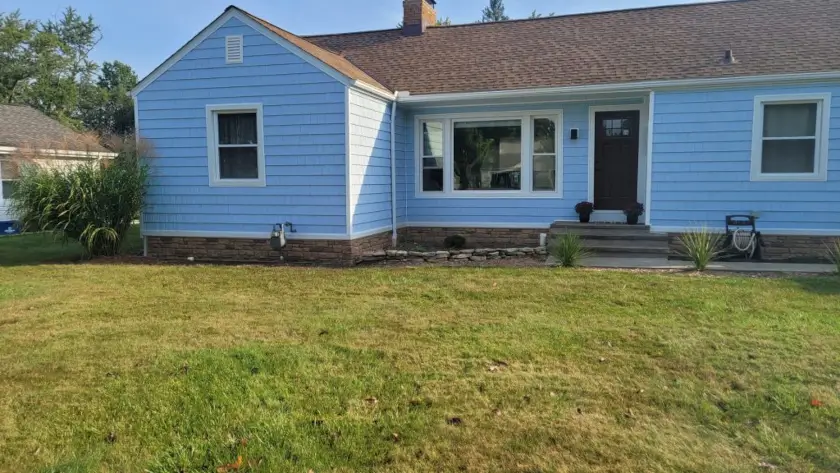 A two-story colonial-style house with light gray vinyl siding is bathed in the cool light of a clear day. A paved driveway leads to the front of the house, which features a covered entryway with columns. Green grass lawns are visible in the foreground on either side of the driveway and a sidewalk runs along the edge of the property. Bare trees are visible in the background, suggesting late autumn or winter. The house has multiple windows with white trim.