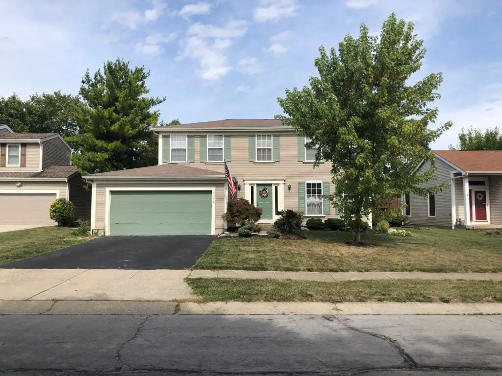 A two-story colonial-style house with light gray vinyl siding is bathed in the cool light of a clear day. A paved driveway leads to the front of the house, which features a covered entryway with columns. Green grass lawns are visible in the foreground on either side of the driveway and a sidewalk runs along the edge of the property. Bare trees are visible in the background, suggesting late autumn or winter. The house has multiple windows with white trim.