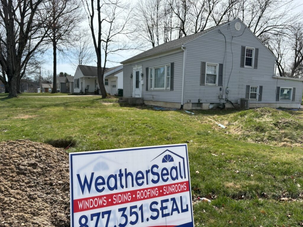 A WeatherSeal yard sign in the grass in front of a light grey residential home.