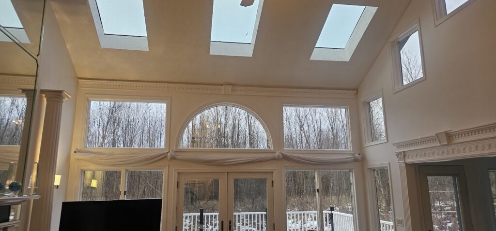 Inside a bright room with tall, angled ceilings and three skylights. A large wall of windows, including a half-circle window in the center, looks out into a wooded area.