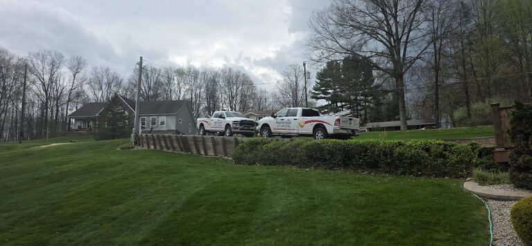 Two white WeatherSeal work trucks parked in a driveway next to a house on a grassy hill with a retaining wall in the front.